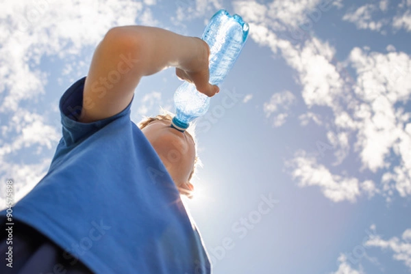 Obraz little boy drinking water from bottle on a hot summer day, thirsty child drinks from water bottle replenish fluids in summer heat wave 

