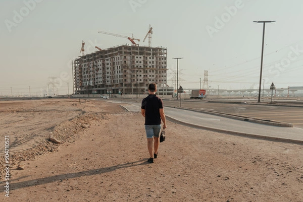 Fototapeta Man walking towards a construction site on a desert road carrying a bag