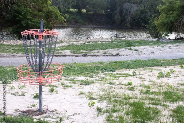 Fototapeta Disc Golf Basket With Chains on Sandy Grass With a River and Trail in the Background