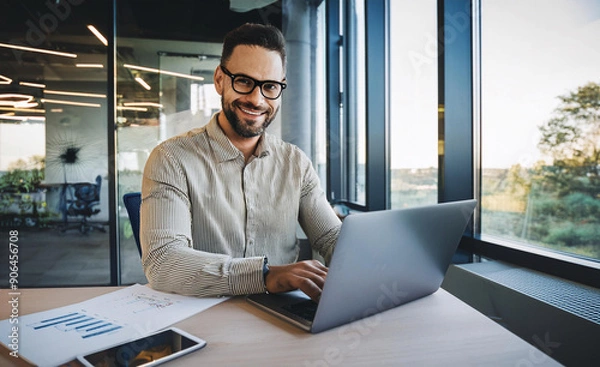 Obraz A smiling young man in a suit sits in the office at a desk and works on a laptop, typing on a keyboard, texting, working with data