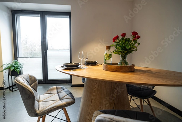 Fototapeta Interior of a Modern furnished Kitchen in a New House. Dining Table and Chairs in Stylish and Functional Space. Detailed kitchen Design. Dining table with plates, glasses and vase of red roses.