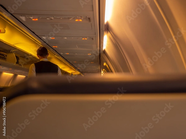 Fototapeta Stewart standing in the aisle of an airplane aircraft cabin. Overhead baggage rack opened and yellow lights turned on. View from behind the passenger seat.