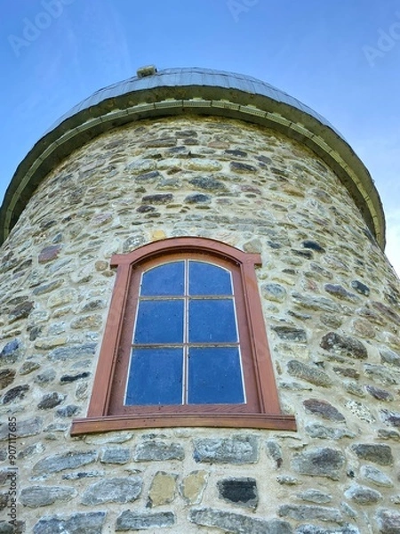 Fototapeta Fieldstone mill with metal roof. Mill tower under a blue sky. Brick tower with old wooden frame window. 