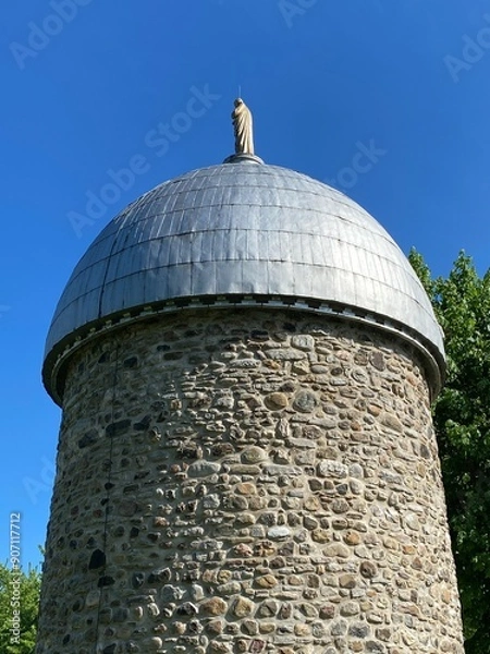 Fototapeta Fieldstone mill with metal roof. Mill tower under a blue sky. Brick tower with old statue of a religious figure from behind on the top.