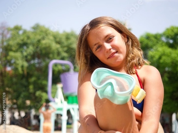 Obraz Close-up portrait of a young caucasian girl holding a snorkel mask at a water park on a sunny day. Green trees and water slides in the background.
