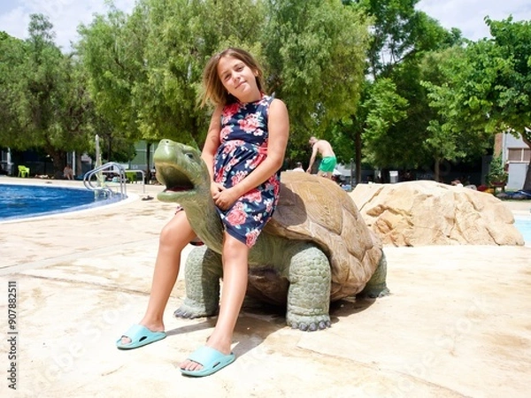 Obraz  Young girl sitting on a large turtle statue by the pool at a water park on a sunny day