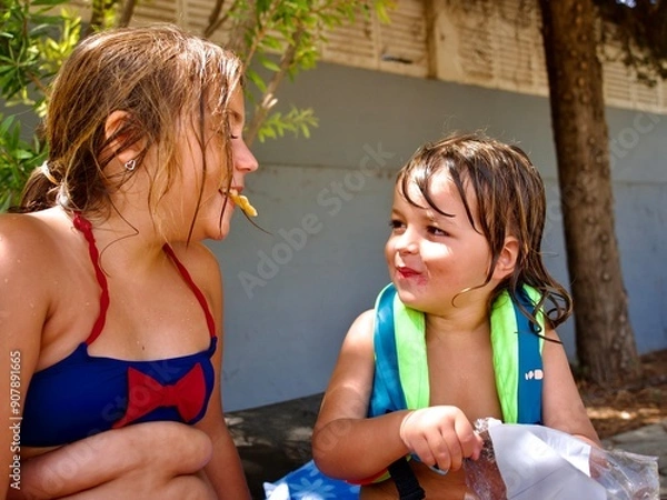 Obraz Close-up portrait of two young caucasian siblings laughing and sharing a snack outside on a sunny summer day. Girl in swimsuit and boy in life jacket.