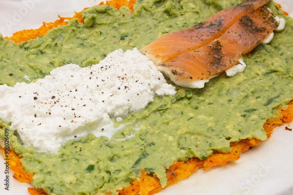 Fototapeta Close-up of a delicious layered dish featuring a base of grated carrots, a middle layer of creamy guacamole, and a topping of white cheese and smoked salmon with herbs on baking paper.