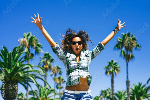 Fototapeta Beautiful funny smiling girl is jumping in palm trees in sunny Barcelona in summer
