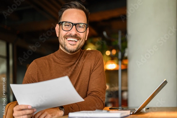 Fototapeta Happy male project manager with document and laptop at desk looking at camera while working in office