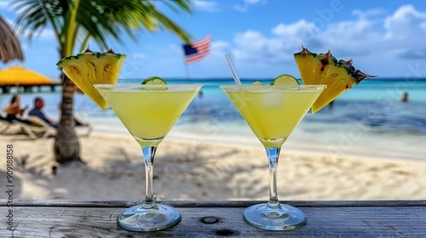 Obraz Two tropical martinis garnished with pineapple slices and lime sit on a beachside table with a vibrant ocean and palm trees in the background - AI Generated Digital Art.