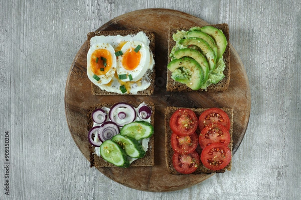 Fototapeta Various sandwiches on a wooden board made of black rye whole grain bread with tomato, avocado, egg and cucumber. Top view