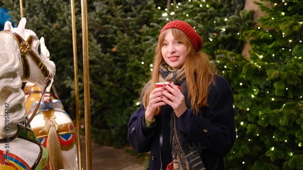 Fototapeta Beautiful young woman with red hair at the Christmas market near carousel in a red beret and coat drinks ,smiling and enjoying hot chocolate from a red cup.Hight quality photo