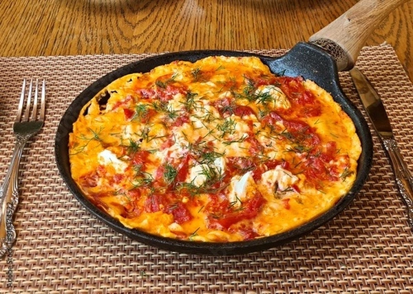 Fototapeta An omelette with tomatoes and fresh herbs is fried in large frying pan over fire