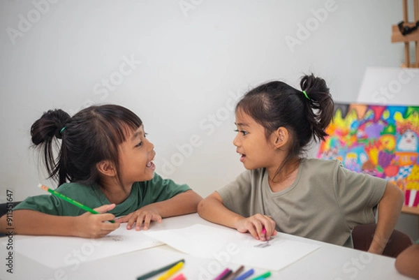 Fototapeta Two young girls are sitting at a table with pencils and coloring books