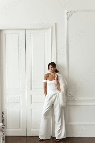 Obraz Beautiful brunette woman dressed in a sleek white suit poses gracefully in a bright, all-white photo studio. Stylish and professional image for business concepts