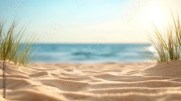 Fototapeta A peaceful view of a sandy beach with gentle waves and green grass under a bright sun, evoking a relaxing coastal atmosphere.