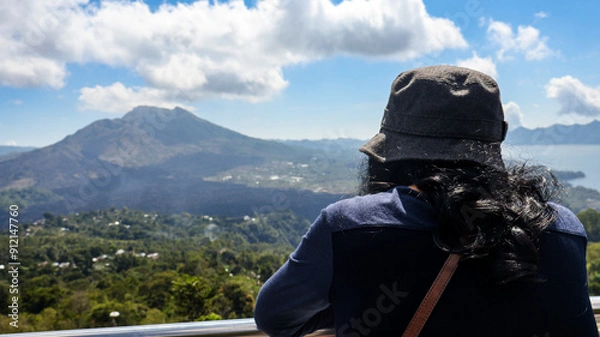 Fototapeta Back view of a female tourist standing on a viewpoint looking at Mount Batur. Volcanic landscape and surrounding greenery are visible under a clear sky, ideal for travel and adventure related content