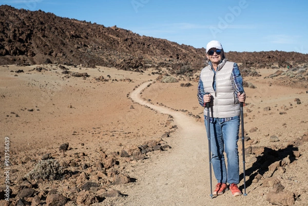 Fototapeta Active cheerful senior woman with backpack walking outdoors with help of poles enjoying nature, freedom and free time. Arid terrain at the base of Teide volcano on the island of Tenerife