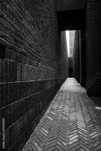 Fototapeta A narrow alleyway between two brick buildings, leading to a brighter area, with a strong contrast between the dark alley and the light at its end.
