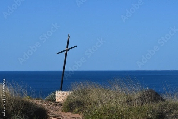 Fototapeta A wooden cross blackened with age and tied together with rope stands on a deserted headland. It rests on sandstone, and is surrounded by wild grasses. It is seen against a blue sea and sky.