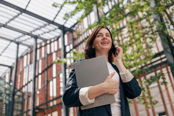 Fototapeta A successful businesswoman in a modern glass building holding a laptop while on a phone call