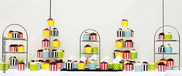 Fototapeta Colourful cup cakes on stainless steel stands shot front on on a white table and white background panorama style