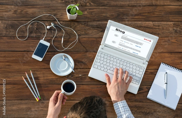 Fototapeta Writer Working on Computer at Wooden Desk Top View of Man Typing on Laptop and Holding Coffee Mug at Warm Natural Wood Table with Electronic Gadgets and Stationery Tools for Every Day Life