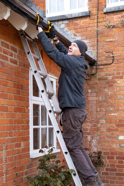 Fototapeta Man on a ladder fixing rain gutter on a UK house