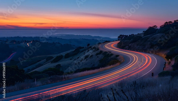 Obraz A long exposure photo of a highway at sunset, with light trails from cars creating streaks across the horizon. The road leads into the distance, with hills in the background, evoking a sense of speed.