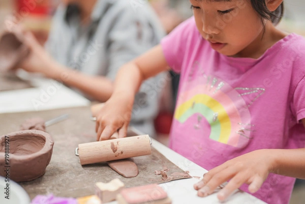 Fototapeta Young girl uses a rolling pin to flatten clay during an art class, Her engagement in the creative process highlights the joy and concentration involved in hands-on learning activities.
