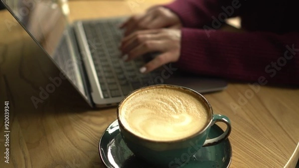Fototapeta Close-up of a cup of coffee with a girl working on a laptop in a cafe in the background.