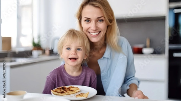 Fototapeta Portrait of the mother and her child in the kitchen, smiling while holding crepes on a white plate with a chocolate spread. 