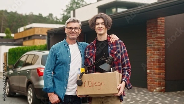 Fototapeta Portrait of father and his son standing outside in yard. Handsome dad hugging his child and proud of his good achievement in college. Satisfied people joyfully smiling while looking on camera.