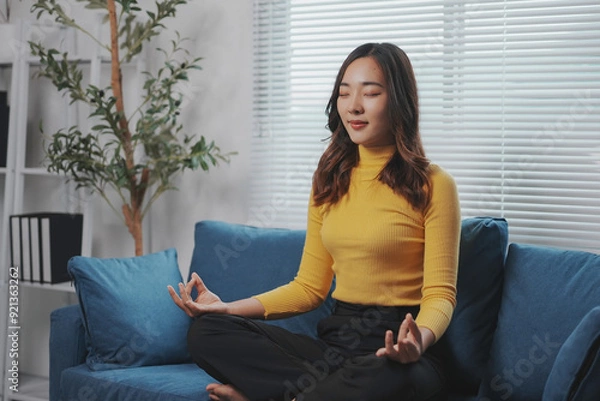 Fototapeta Young asian woman is practicing mindfulness with her eyes closed while sitting in the lotus position on a sofa at home