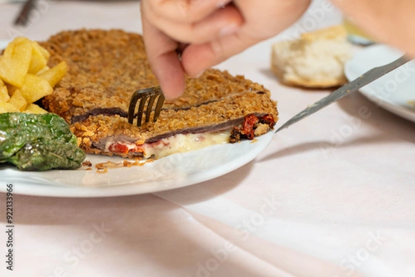Fototapeta Hands split a cachopo, beef fillet, with serrano ham, red bell pepper and cheese inside.