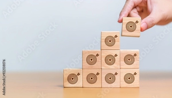 Obraz Hand holding wooden block with target aiming dart board and arrow up on table background. Business achievement objective goal and project progress growth value processing marketing financial concept