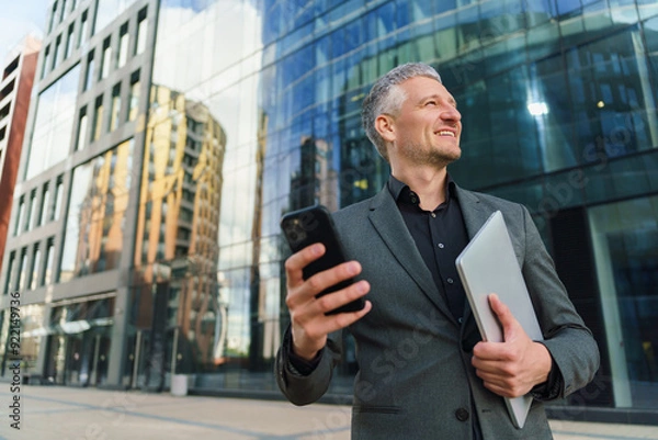 Fototapeta A confident businessman in a gray suit holds a smartphone and a tablet, smiling while looking up in a modern urban setting.
