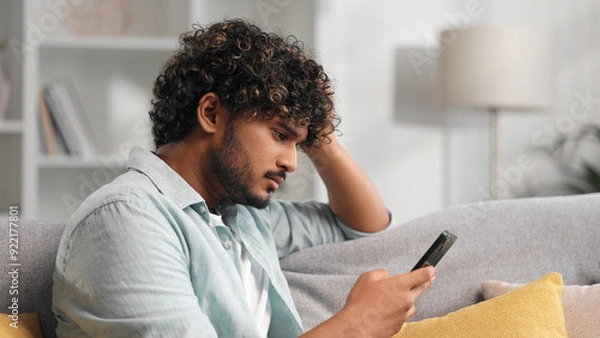 Obraz Worried young Indian boy sitting on sofa looking at her phone and does not understand why it is not working. The angry student in glasses does not believe the bad news he saw.