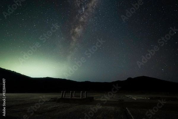 Fototapeta The Milky Way galaxy above a silhouette of mountains with part of a ruined tracking station in the foreground. You can see a glow of light over the mountains in the distance with a starry sky above.