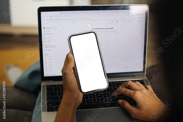 Fototapeta Hand Holding Smartphone With Blank White Screen For Application Mockup While Using Laptop
