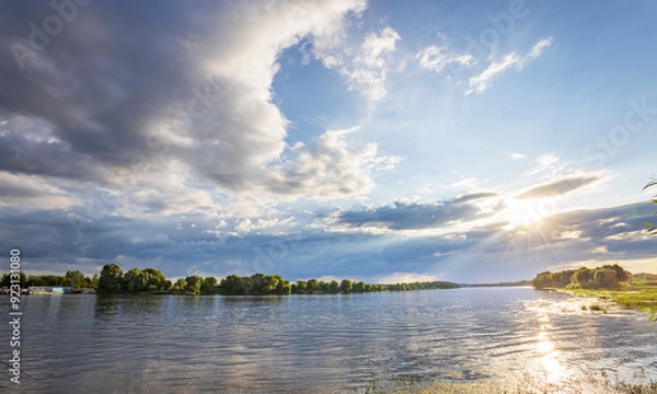 Fototapeta A serene river flows under a sky filled with fluffy clouds. The golden light filters through, casting a glow on the calm water. Trees on the opposite bank complete the beautiful scene