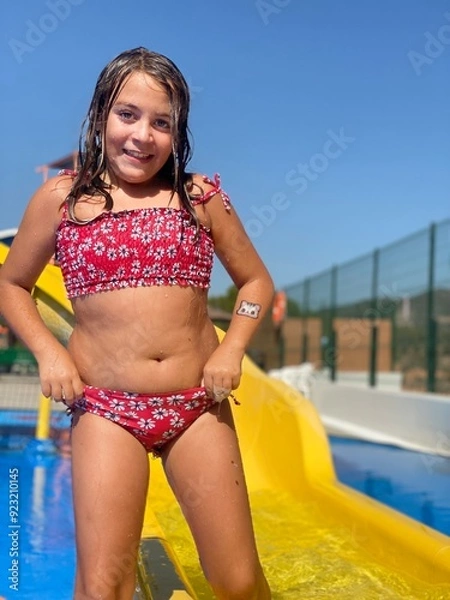 Obraz A young girl in a red swimsuit smiling at the camera after a fun slide at an outdoor water park, with clear blue skies in the background.