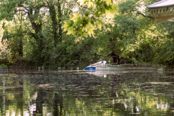 Fototapeta In a magical corner, a boat floats on a mirror-like pond. The cabin among trees and the sky’s reflection create a peaceful haven, where time stands still and nature whispers serenity