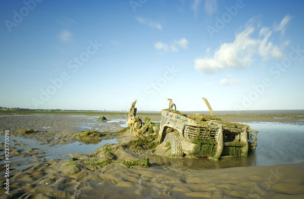 Fototapeta An abandoned half buried rusting and barnacle encrusted 4x4 left to fall apart on a wet sandy beach. A long daytime exposure to blur the water and the clouds.