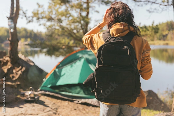 Fototapeta A woman with a backpack looks out over a forest lake. In front of her is a green tent and a beautiful view of peaceful nature at dawn.
