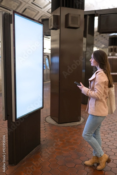 Obraz Metro station, a young woman looking at a wall map for directions, holding a cellphone with copy space.