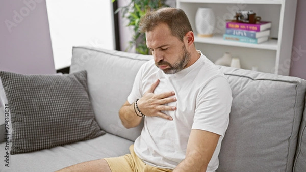 Obraz Mature hispanic man with grey hair feeling chest pain in a home interior with sofa background.