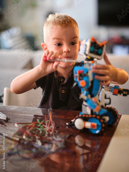 Obraz A young child is depicted using a tool to fix a colorful robot toy, emphasizing creativity and focus as part of a hands-on learning experience indoors.