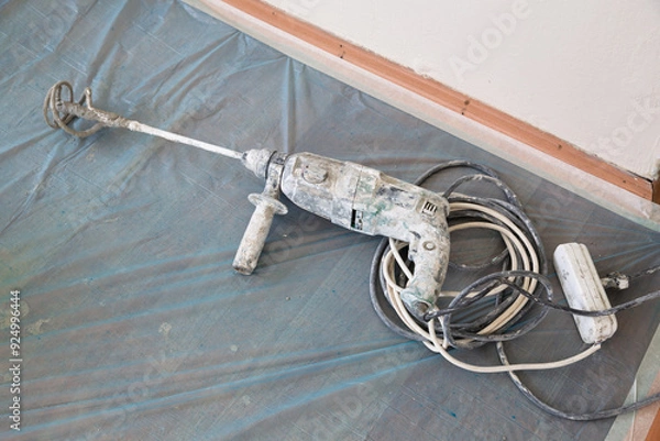 Fototapeta Large hammer drill with mixer bit, power cable and three-way socket. A blue plastic cleaning tarpaulin is laid on the floor.